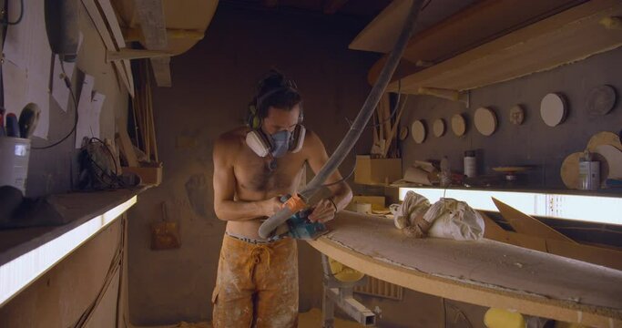 Gimbal shot of shirtless shaper with electric planer working on surfboard
