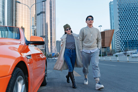 a Fashionable young couple with sunglasses and walking beside an orange luxury sports car