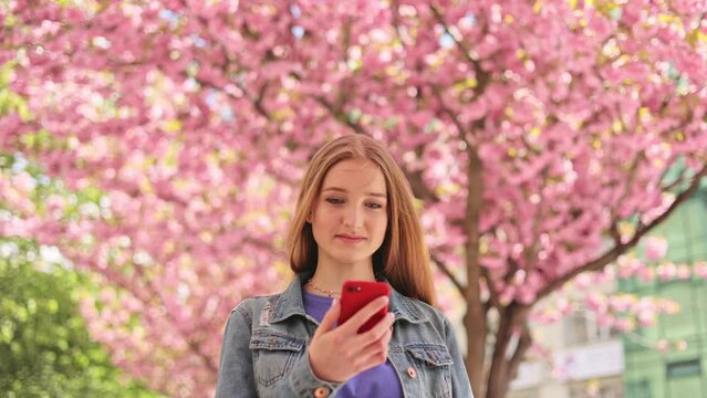 Portrait shot of beautiful young woman look at smartphone and smiling on blurred background in the city street. Sacura tree. Caucasian beautiful female walking around sakura trees and texting on phone
