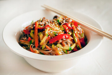Traditional Korean starch noodles chapche with vegetables and beef in a white round plate on a gray background, chopsticks lay on a plate with noodles. side view, close-up.