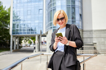 Side view of concentrated woman in stylish outfit text messaging on mobile phone while walking along near contemporary building