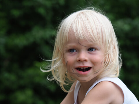 Portrait face baby boy blonde hair. A dirty child against the background of green trees. Happy cute adorable kid laughs fervently