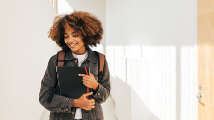 Cheerful student with curly hair standing in hallway. Girl holding notebook and looking down.