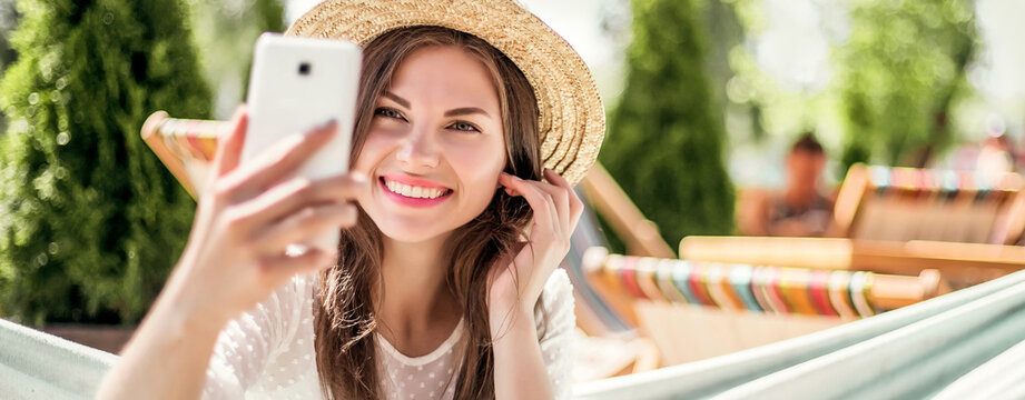 Fashionable young beautiful woman smiling, taking selfie on mobile phone, making video conference calls while on vacation