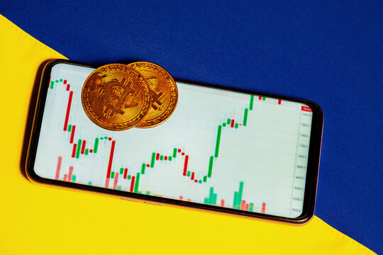 Bitcoin and mobile phone with cryptocurrency market graph on yellow blue Ukrainian flag close up. Cryptocurrency background for virtual money.