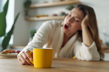 Young woman in bathrobe yawning with cup of coffee while sitting at the kitchen counter at home - obrazy, fototapety, plakaty
