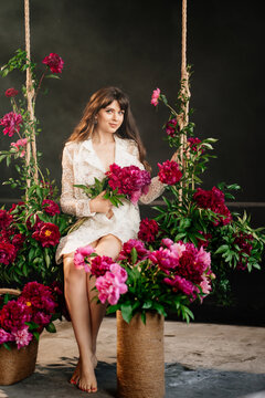 a beautiful young woman in a white dress sits on a swing in peony flowers. 