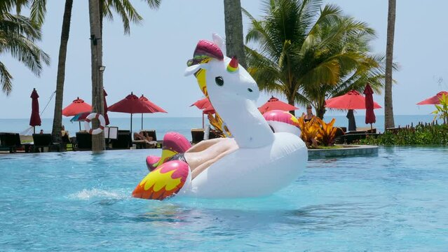 Happy funny man unsuccessfully jump onto floating inflatable unicorn in swimming pool in tropical hotel to ride. Smiling male tourist have fun with giant toy in water. Summer vacation, holiday concept