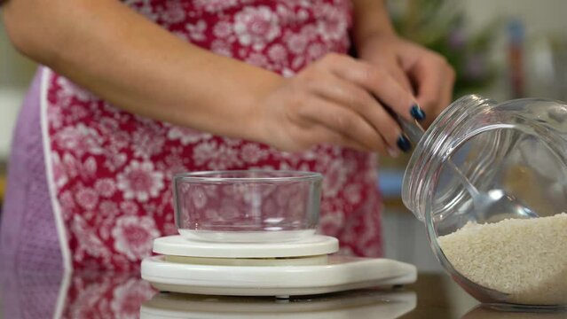 White rice on kitchen scale. Female's hand is pouring jasmine rice in transparent bowl on electronic scale. Weighing products on digital scale.  Weighing loose cereals rice millet. 