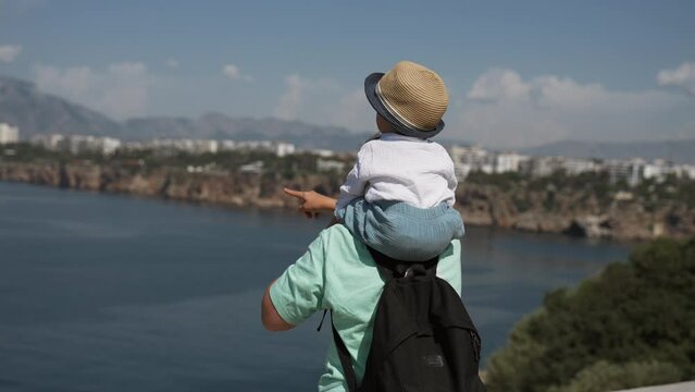 Mom shows toddler boy in sun protective hat sitting on shoulders beautiful views of sea. Woman and child walk on waterfront in coastal town slow motion