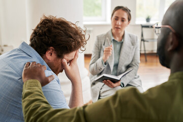 Worried young male patient keeping hand on head while black man comforting him during psychological...