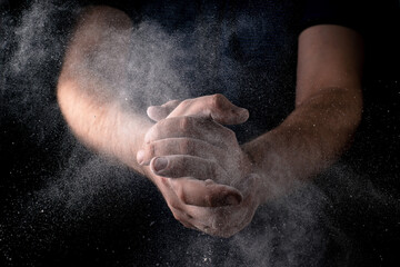 A man claps his hands with scattering flour against a dark background. Culinary theme.