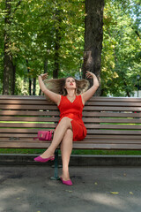 Attractive blonde woman on bench in park. Young woman in red dress relaxes in park on summer day. Vertical frame.