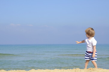 A little boy stands on the beach and looks beyond the horizon. A child in striped shorts and a white T-shirt hugs the sea, the ocean.