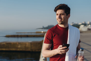 Young strong sporty athletic fit sportsman man in sports clothes towel hold use mobile cell phone water bottle warm up training at sunrise sun dawn over sea beach outdoor on pier seaside in morning.