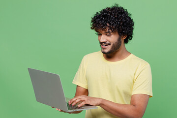 Young smiling happy Indian man 20s in basic yellow t-shirt hold use work on laptop pc computer chatting online isolated on plain pastel light green background studio portrait People lifestyle concept - obrazy, fototapety, plakaty