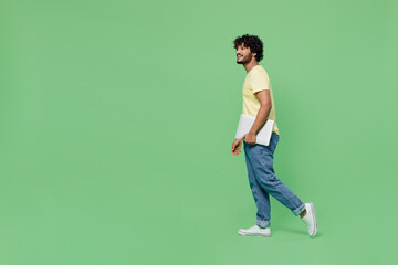 Fototapeta na wymiar Full body side view young smiling happy fun Indian man 20s in yellow t-shirt hold closed laptop pc computer look aside on workspace mock up walk isolated on plain pastel light green background studio.
