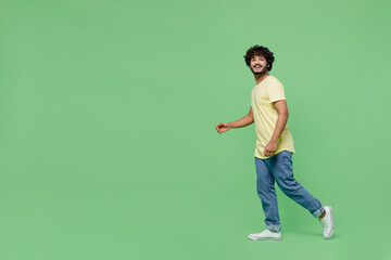 Fototapeta na wymiar Full body young smiling happy Indian man 20s wearing basic yellow t-shirt walk going strolling look camera isolated on plain pastel light green background studio portrait. People lifestyle concept