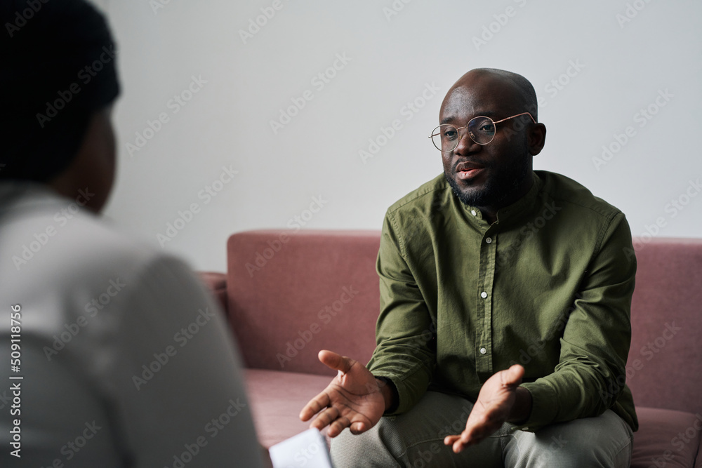 Wall mural young black man in casualwear sitting on couch and explaining his emotions and worries during talk w