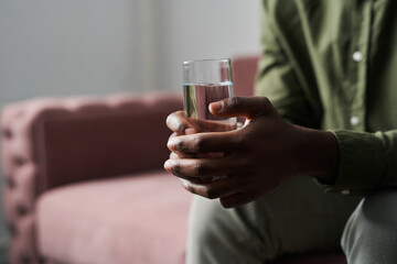 Hands of young troubled African American man holding glass of water while sitting on couch in office of his psychoalyst or psychologist