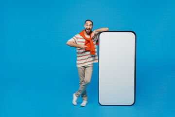 Full size young man 20s in orange striped t-shirt point index finger on big blank screen mobile cell phone with workspace copy space mockup area show thumb up gesture isolated on plain blue background