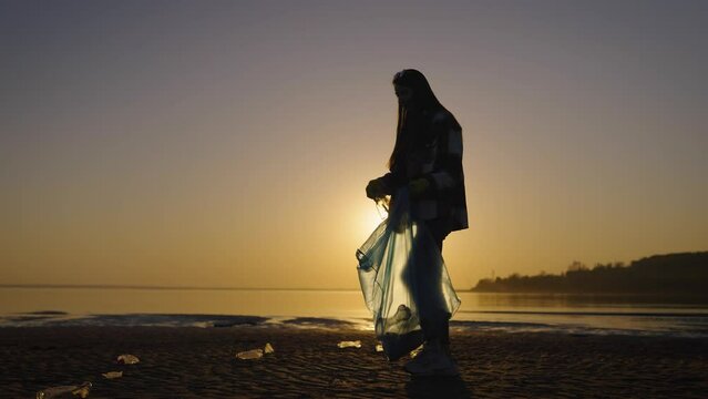 Silhouette of a woman with a trash bag at sunset. A volunteer collects and sorts waste on the beach.
