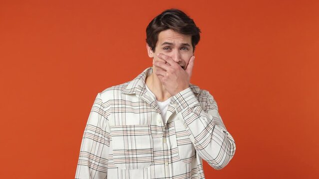 Scared frightened shocked young brunette man 20s years old wears white shirt look camera covering hiding face with hands peep through fingers isolated on plain orange wall background studio portrait
