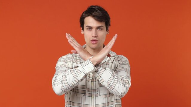 Serious strict dissatisfied sad severe young brunette man 20s years old wears white shirt say no hold palm folded crossed hands in stop gesture isolated on plain orange wall background studio portrait