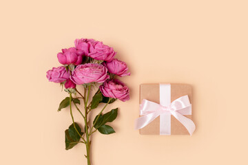 Gift box with a tied bow and bouquet of pink rose flowers on a beige background. Congratulation concept with copyspace.