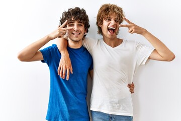 Young gay couple standing together over isolated background doing peace symbol with fingers over face, smiling cheerful showing victory