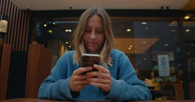 Attractive young woman with light hair wearing blue sweater, sitting inside cosy cafe, using smartphone, smiling, typing, texting, chatting with friends on social media, scrolling news on mobile phone