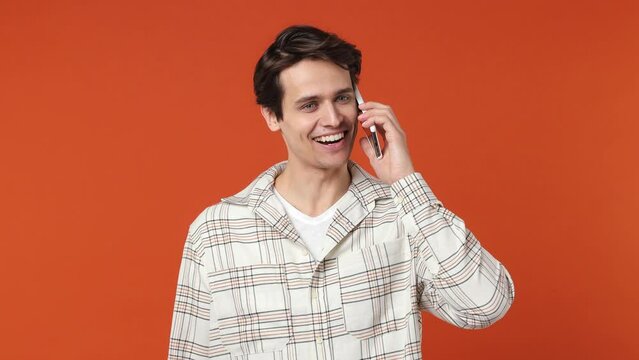 Happy calm smiling vivid young brunet man 20s years old wears white shirt hold use talk on mobile cell phone conducting pleasant conversation isolated on plain orange wall background studio portrait