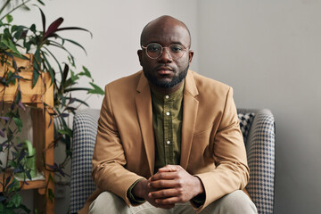 Young serious well-dressed male psychotherapist in eyeglasses sitting in armchair in front of...