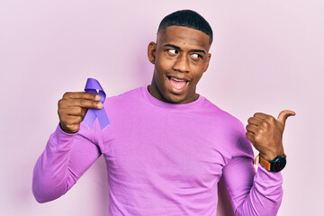 Young black man holding purple ribbon awareness pointing thumb up to the side smiling happy with...