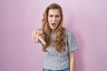 Beautiful blonde woman standing over pink background pointing displeased and frustrated to the...