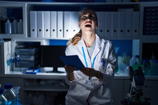 Beautiful blonde woman working at scientist laboratory late at night angry and mad screaming frustrated and furious, shouting with anger. rage and aggressive concept.