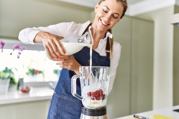 Young blonde woman smiling confident pouring milk on blender at kitchen