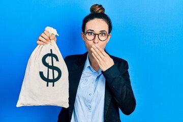 Young hispanic girl wearing business clothes holding dollars bag covering mouth with hand, shocked...
