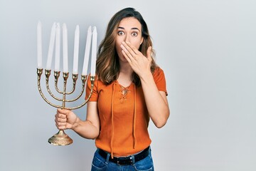 Young hispanic girl holding menorah hanukkah jewish candle covering mouth with hand, shocked and...