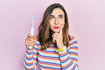 Obraz premium Young hispanic girl holding electric toothbrush serious face thinking about question with hand on chin, thoughtful about confusing idea