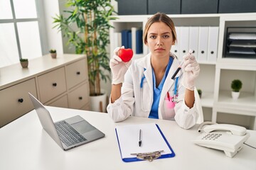 Young blonde woman wearing doctor uniform holding blood and heart relaxed with serious expression on face. simple and natural looking at the camera.