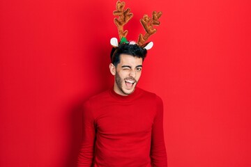 Young hispanic man wearing cute christmas reindeer horns winking looking at the camera with sexy...