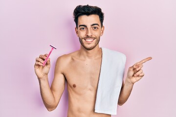Young hispanic man standing shirtless holding razor smiling happy pointing with hand and finger to the side