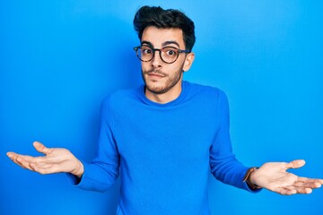Young hispanic man wearing casual clothes and glasses clueless and confused expression with arms...