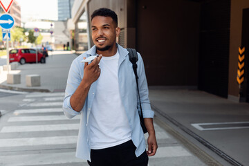 Handsome african american guy recording voice message on cellphone while walking on city street,...