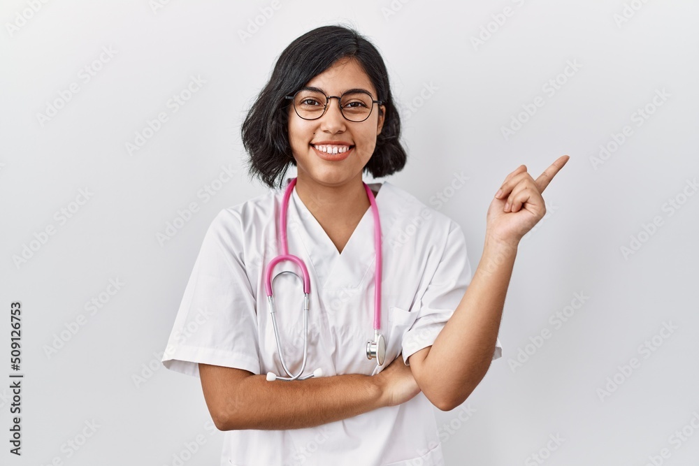 Sticker Young hispanic doctor woman wearing stethoscope over isolated background with a big smile on face, pointing with hand and finger to the side looking at the camera.