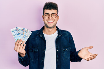 Young hispanic man holding peruvian sol banknotes celebrating achievement with happy smile and winner expression with raised hand - obrazy, fototapety, plakaty