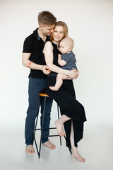 Portrait of young happy family in dark clothes with plump cherubic baby infant toddler standing on white background.