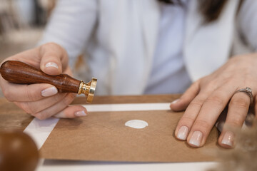 Close-up of hands with rings of unrecognizable woman making wax seal with stamp on brown greeting invitation on desk.