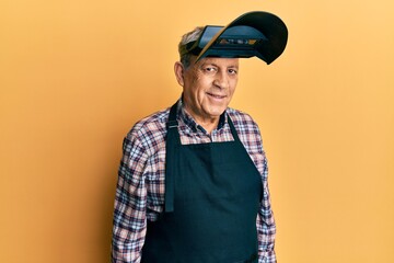 Handsome senior man with grey hair wearing welding protection mask with a happy and cool smile on...